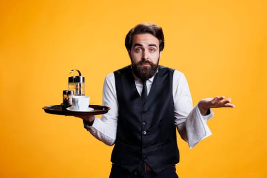 Clueless worker with platter in hand raising shoulders and expressing doubtful feelings, posing against yellow background. Professional waiter acting uncertain and shrugging on camera.