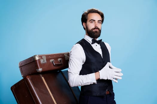 Professional hotel porter wears gloves posing over blue background, preparing to carry trolley bags and help guests. Young employee working as bellhop, tourism industry concept.