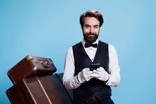 Hotel porter scrolls on smartphone, using social media and texting messages while he poses in studio with trolley bags. Doorman bellhop in suit browsing online page on phone app.