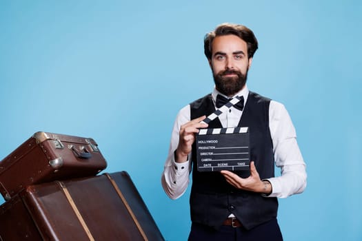 Doorkeeper clapping filmslate on camera, saying action to cut scene for fun against blue background. Classy hotel bellboy pretending to work in movie production using clapperboard.