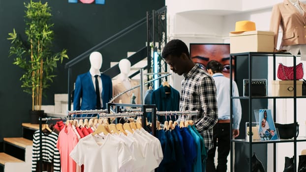 African american shopper checking basic shirt, analyzing merchandise fabric in clothing store. Shopaholic customer shopping for fashionable clothes in modern boutique. Fashion concept