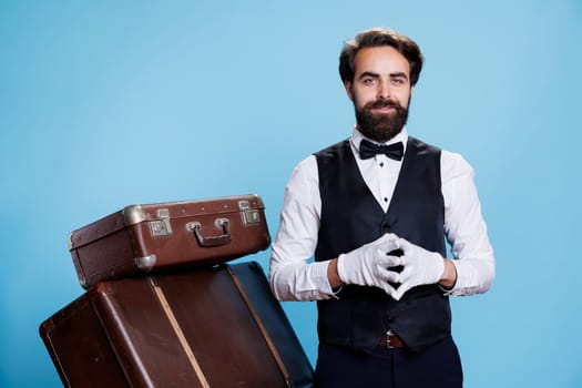 Young man hotel concierge with gloves helping guests to carry luggage and trolley bags, smiling with confidence in studio. Luxury doorman bellhop working in hospitality and tourism industry.