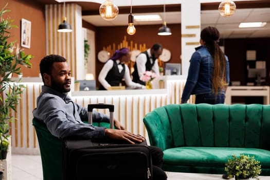 Young African American man traveler waiting for hotel check-in, sitting in armchair with luggage beside, resting in cozy comfortable lounge area. Black guy businessman with baggage in lobby