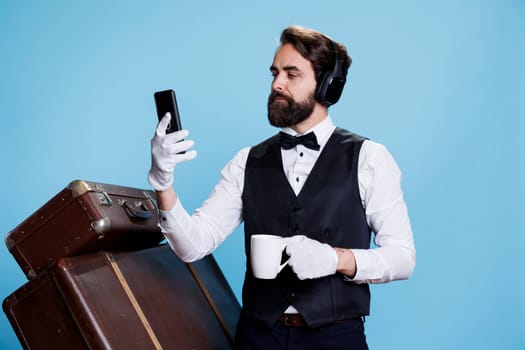 Modern bellhop checks messages and holding cup of coffee in studio, enjoying break with music on headset. Young man with professional hotel industry job texting and drinking cold brew.