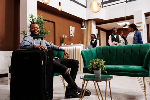 Happy smiling African American man guest sitting in hotel lobby with suitcase awaiting for room. Positive millennial black guy tourist resting in armchair while waiting for check-in time