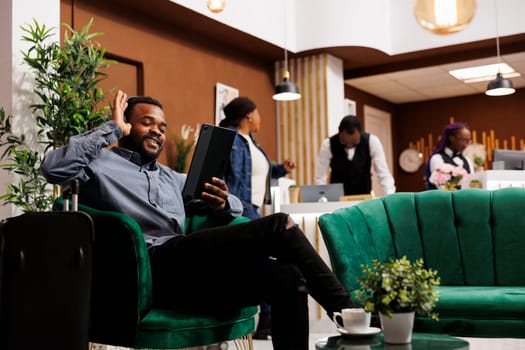 Happy smiling young African American man traveler sitting in hotel lobby having video call while waiting for check-in. Tourist holding digital tablet, waving hand greeting friend looking at camera