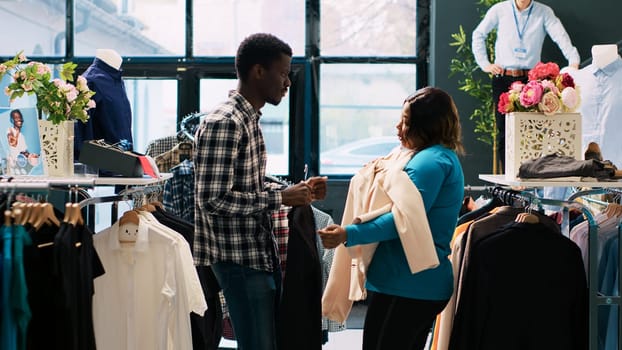 Playful couple fooling around in modern boutique, playing with stylish merchandise during shopping session. African american clients buying for fashionable items in clothing store. Fashion concept