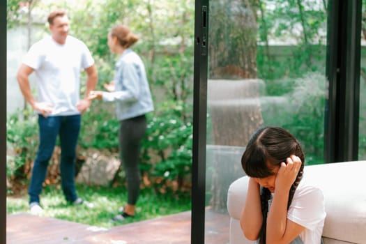 Stressed and unhappy young girl huddle in corner, cover her ears blocking sound of her parent arguing in background. Domestic violence at home and traumatic childhood develop to depression. Synchronos