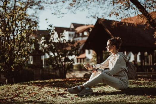 Young fashionable teenage girl with smartphone in park in autumn sitting at smiling. Trendy young woman in fall in park texting. Retouched, vibrant colors. Beautiful blonde teenage girl wearing casual modern autumn outfit sitting in park in autumn. Retouched, vibrant colors, brownish tones.