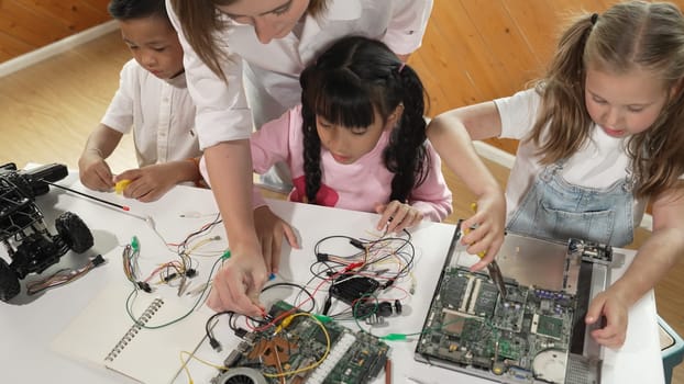 Top view of caucasian teacher teaching diverse students about electronic board. Multicultural children learn about digital electrical tool and fixing motherboard by using chips and wires. Erudition.