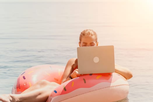 Woman freelancer works on laptop swimming in sea on pink inflatable ring. Pretty lady typing on computer while floating in the sea on inflatable donut at sunset. Freelance, remote work on vacation