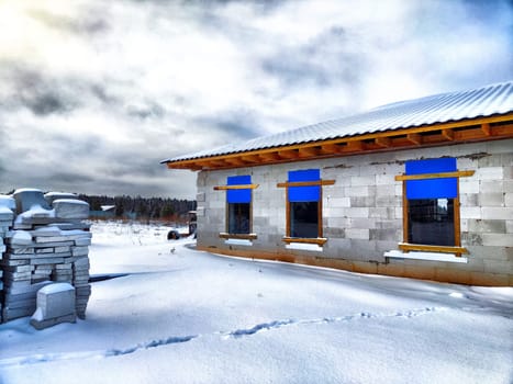 An unfinished house and building materials in the snow. Building a new house in winter. An unfinished house with building materials surrounded by snow