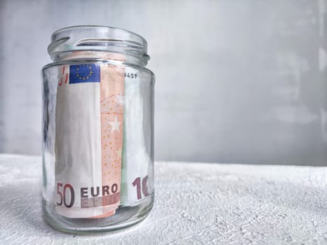 Glass Jar Filled With Euro Banknotes on Textured White Surface. Euro currency in clear jar as background. Concept of public distrust of banks and keeping money at home