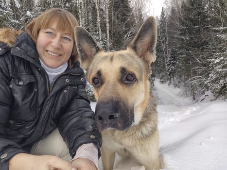 Adult girl with shepherd dog taking selfies in winter forest. Middle aged woman and big shepherd dog on nature in cold day. Friendship, love, communication, fun, hugs