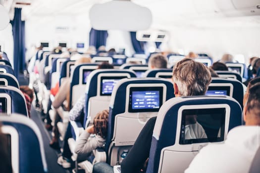 Interior of airplane with passengers on seats and stewardess in uniform walking the aisle, serving people. Commercial economy flight service concept