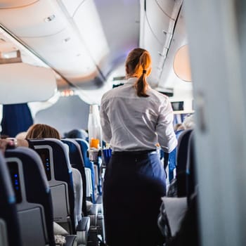 Interior of airplane with passengers on seats and stewardess in uniform walking the aisle, serving people. Commercial economy flight service concept