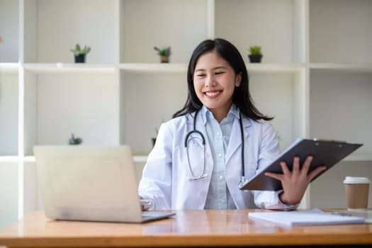 young asian lady doctor in white medical uniform with stethoscope using computer laptop talking video conference call with patient at desk in health clinic or hospital..