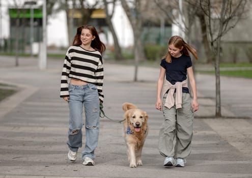 Two Sisters With A Golden Retriever Are Walking On The Street In Summer