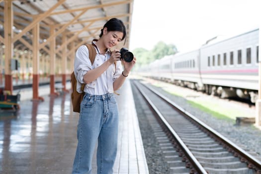 Young Asian woman holds a camera and takes photos of a train while waiting for the train at the train station to travel..