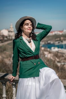 Woman walks around the city, lifestyle. A young beautiful woman in a green jacket, white skirt and hat is sitting on a white fence with balusters overlooking the sea bay and the city