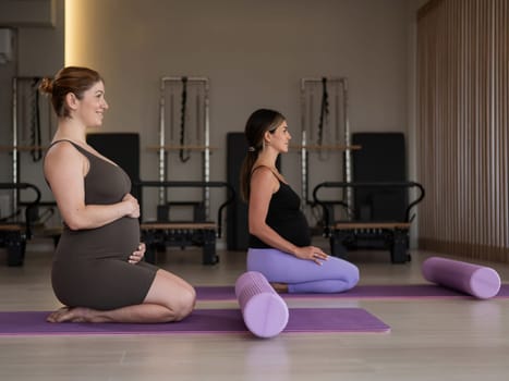 Two pregnant women are sitting on a yoga mat. Prenatal yoga