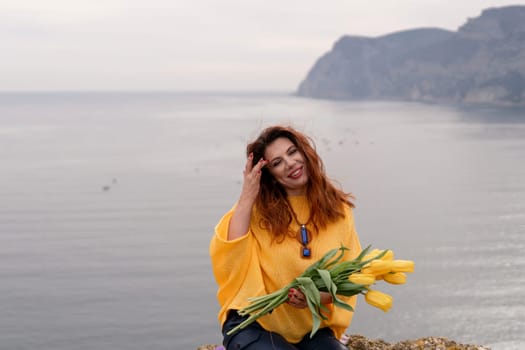 Portrait happy woman woman with long hair against a background of mountains and sea. Holding a bouquet of yellow tulips in her hands, wearing a yellow sweater.