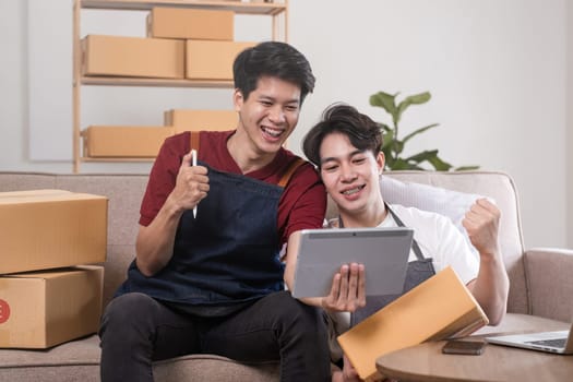 A gay couple running a small business takes order on a laptop and sells product online together in a room full of boxes..
