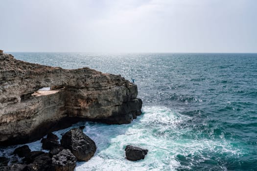 The ocean is calm and the sky is cloudy. The water is a deep blue color. The rocks are jagged and the waves are small