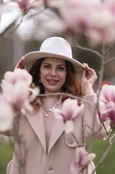 Woman magnolia flowers, surrounded by blossoming trees, hair down, white hat, wearing a light coat. Captured during spring, showcasing natural beauty and seasonal change