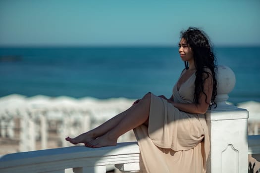 A woman in a tan dress is sitting on a white railing overlooking the ocean. The scene is serene and peaceful, with the woman enjoying the view and the calming sound of the waves