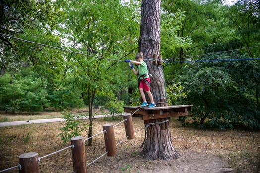 Strong excited young boy playing outdoors in rope park. Caucasian child dressed in casual clothes and sneakers at warm sunny day. Active leisure time with children concept.