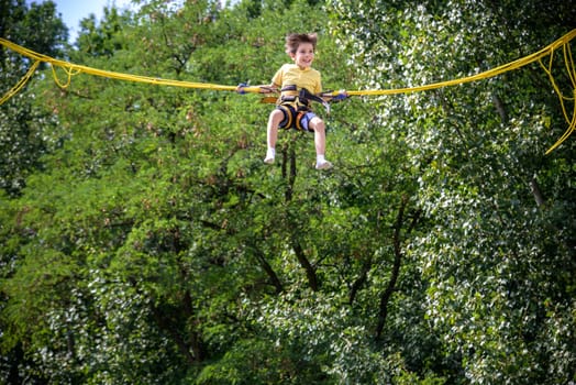 The boy is jumping on a bungee trampoline. A child with insurance and stretchable rubber bands hangs against the sky. The concept of happy childhood and games in the amusement park.