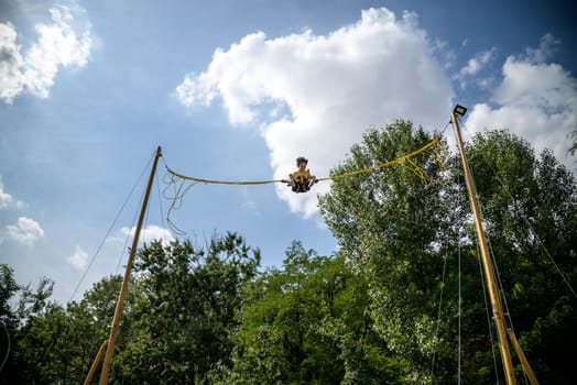 The boy is jumping on a bungee trampoline. A child with insurance and stretchable rubber bands hangs against the sky. The concept of happy childhood and games in the amusement park.