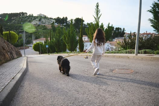 Full length portrait of European cute little child girl in sportswear, walking her dog on leash on the street. People and animals. Playing pets concept