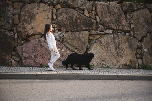 Full length portrait of European cute little child girl in sportswear, walking her dog on leash on the city street. People and animals. Playing pets concept