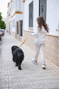 Full length portrait of European cute little child girl in sportswear, walking her dog, a purebred black cocker spaniel on leash on the street. People and animals. Playing pets concept. Rear view