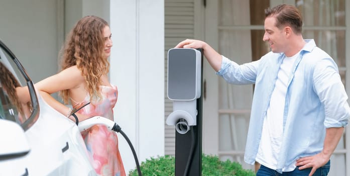 Happy and lovely couple with eco-friendly conscious recharging electric vehicle from EV home charging station. EV car technology for home utilization to future sustainability. Panorama Synchronos