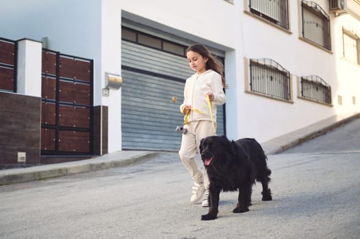 Full length authentic portrait of a lovely Caucasian child pretty girl in white sports wear, smiling enjoying a walk with her cute companion pet, a black purebred cocker spaniel dog in the city street