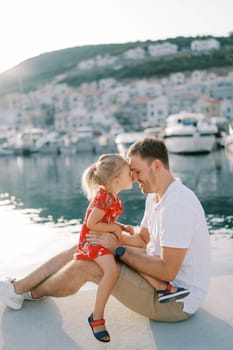 Little girl sits on her dad lap with her nose touching him on the boardwalk by the sea. High quality photo