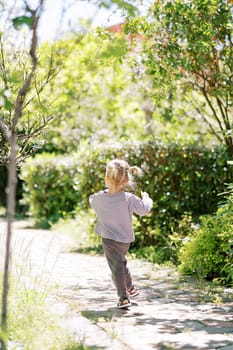 Little girl runs along a path in a green park, waving her arms. Back view. High quality photo