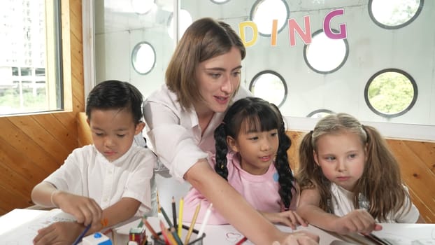 Teacher clicking at laptop while diverse student looking at screen. Children excited in presentation while drawing or writing art books at table with toys and color pencil placed on table. Erudition.