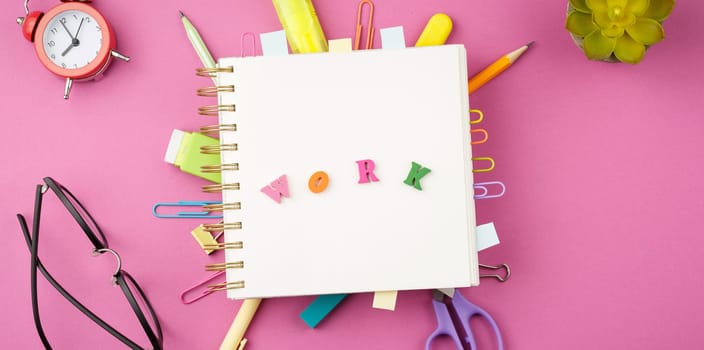 Open spiral notebook with bookmarks from paper clips and leaves for notes, pencil, scissors, binders, alarm clock, glasses on a pink background. Top view. Stationery concept. Office tools.