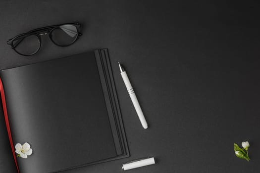 Open notebook with red bookmark and black sheets, white pen, glasses and white flowers on dark isolated background flat lay.