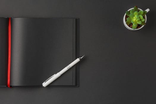 Black notebook with red ribbon bookmark and white pen on it and white cup for green ornamental plant on a dark background, flat lay.