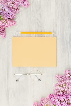 Craft notebook with pencil, glasses and lilac flowers on a white wooden background. Top view.