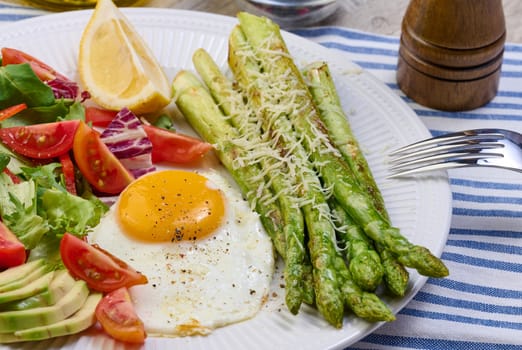 Round plate with cooked asparagus, fried egg, avocado and fresh vegetable salad on the table, top view