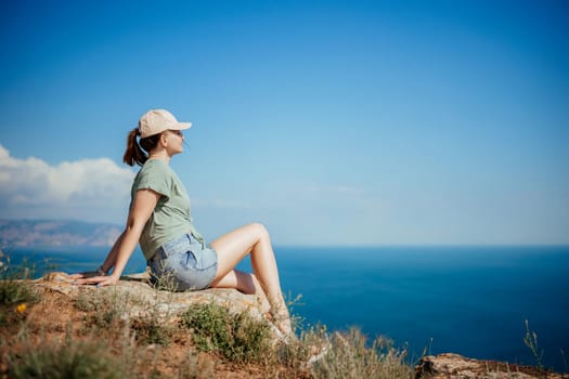 A woman is sitting on a rock overlooking the ocean. She is wearing a green shirt and blue shorts. The sky is clear and blue, and the ocean is calm. The woman is enjoying the view