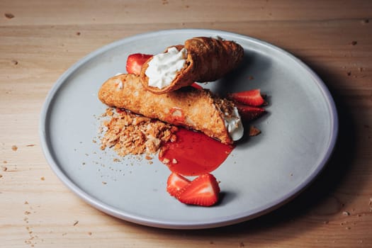 A cannoli filled with cream, accompanied by strawberries and a crumbled topping, served on a stylish gray plate.