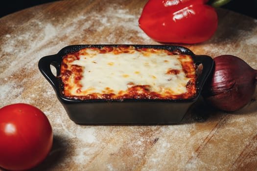 Baked Lasagna in a BlFreshly baked lasagna with melted cheese in a ceramic dish, accompanied by tomatoes and a red bell pepper.ck Ceramic Dish Surrounded by Fresh Vegetables. High quality photo