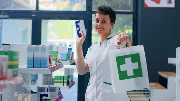 Portrait of happy druggist in apothecary excited to provide ailments relieve products to clients. Upbeat wellness expert holding prescription pills box and shopping bag in drugstore